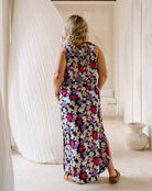 A woman viewed from behind wearing a sleeveless, floor-length floral maxi dress with a multicolor pattern of pink, purple, white, and green flowers on a dark background. She is standing in a brightly lit, neutral-toned room with draped white fabric and a ceramic vase in the background.