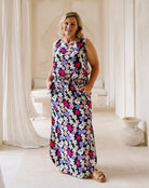 A woman modeling a sleeveless, floor-length maxi dress with a colorful floral pattern on a dark background, standing in a bright, neutral-toned indoor setting.