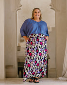 A woman modeling a two-piece outfit consisting of a blue short-sleeve blouse and a black maxi skirt with a vibrant pink, purple, and white floral pattern, standing in front of a beige arched doorway.