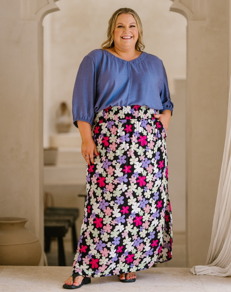 A woman modeling a two-piece outfit consisting of a dusty blue short-sleeve blouse and a long black maxi skirt with a vibrant pink, purple, and white floral pattern, standing in a warm, neutral-toned indoor setting.