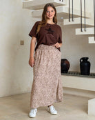 Woman wearing a brown t-shirt and patterned skirt standing in a modern interior setting.