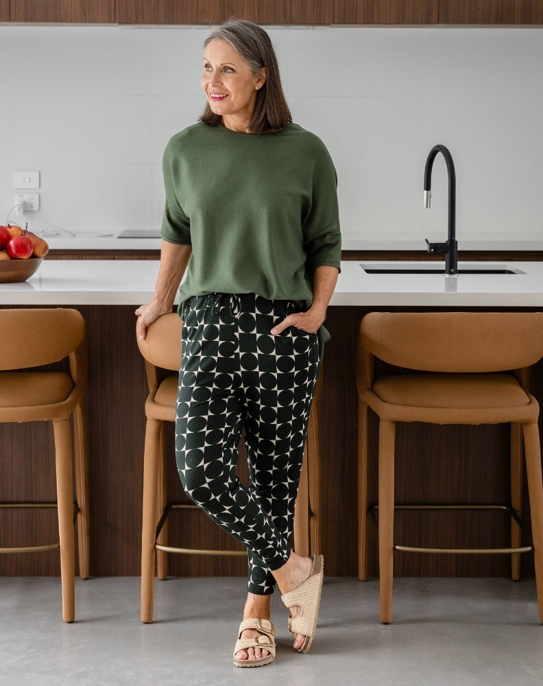 Woman standing in a modern kitchen wearing a green sweater and patterned pants.