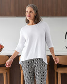 Woman standing in a kitchen wearing a white top and patterned pants.