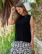 Woman wearing a black sleeveless top and patterned skirt standing outdoors with greenery and a stone wall in the background.