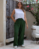 Woman wearing a white top and green pants standing in an outdoor setting with a wooden door and plant in the background.