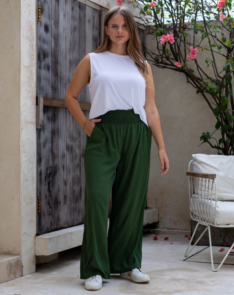 Woman wearing a white top and green pants standing in an outdoor setting with a wooden door and plant in the background.