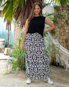 Woman wearing a black top and patterned skirt standing outdoors with plants and a stone wall in the background.