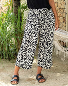 Person wearing black and white patterned pants standing outdoors with greenery and a stone wall in the background.