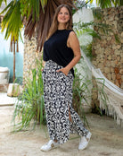 Woman wearing a black top and patterned pants standing in a tropical setting with plants and a stone wall.