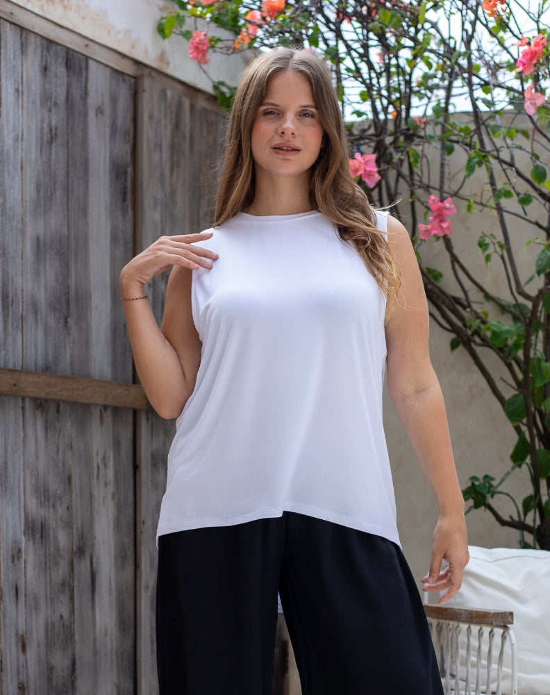 Woman wearing a white sleeveless top and black pants standing outdoors with a wooden fence and plants in the background.