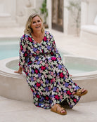 A woman wearing a long-sleeve maxi dress with a colorful floral print sits on the edge of an outdoor pool. The dress has a black base with pink, purple, white, and green flowers, and she is wearing brown strappy sandals.