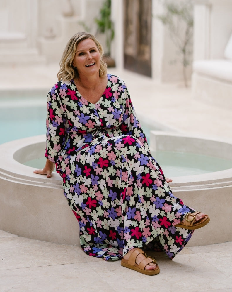A woman wearing a long-sleeve maxi dress with a colorful floral print sits on the edge of an outdoor pool. The dress has a black base with pink, purple, white, and green flowers, and she is wearing brown strappy sandals.