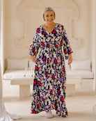 A woman wearing a long-sleeved maxi dress with a colorful floral print, standing in a bright, neutral-toned indoor setting with a stone bench and carved wall behind her.