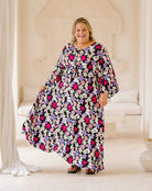 A woman modeling a loose, long kaftan dress with a vibrant floral print in pink, purple, and white on a black background, set in a bright, neutral-toned indoor studio environment.