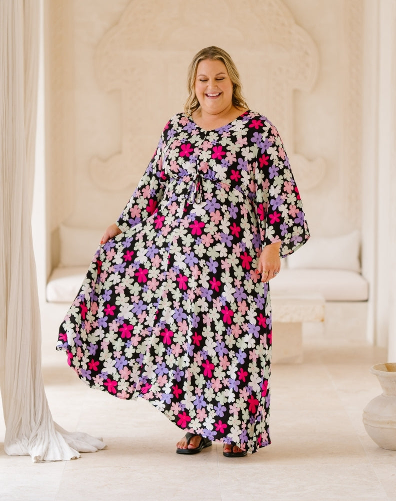 A woman modeling a loose, long kaftan dress with a vibrant floral print in pink, purple, and white on a black background, set in a bright, neutral-toned indoor studio environment.