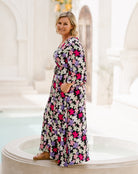 A woman wearing a long-sleeved, knee-length maxi dress with a vibrant floral print in pink, purple, white, and black, standing on a white stone terrace in a bright, sunlit architectural setting.