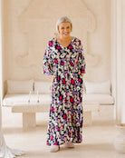 A smiling woman stands in a bright, cream-colored indoor setting wearing a long-sleeved maxi dress with a dense floral print. The dress features black, pink, purple, and white flowers.