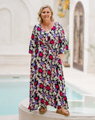 A woman wearing a long-sleeve, V-neck maxi dress with a colorful floral print, standing in front of a white architectural background with a pool.