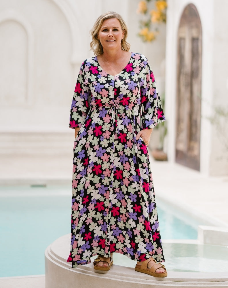 A woman wearing a long-sleeve, V-neck maxi dress with a colorful floral print, standing in front of a white architectural background with a pool.
