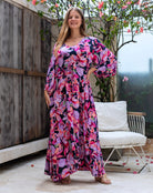 Woman wearing a colorful floral dress standing outdoors near a wooden wall and white chair.