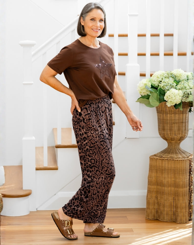 Woman in brown shirt and leopard print pants standing in a home setting with flowers in the background.