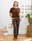 Woman in brown shirt and leopard print pants standing in a home setting with a staircase and flowers.